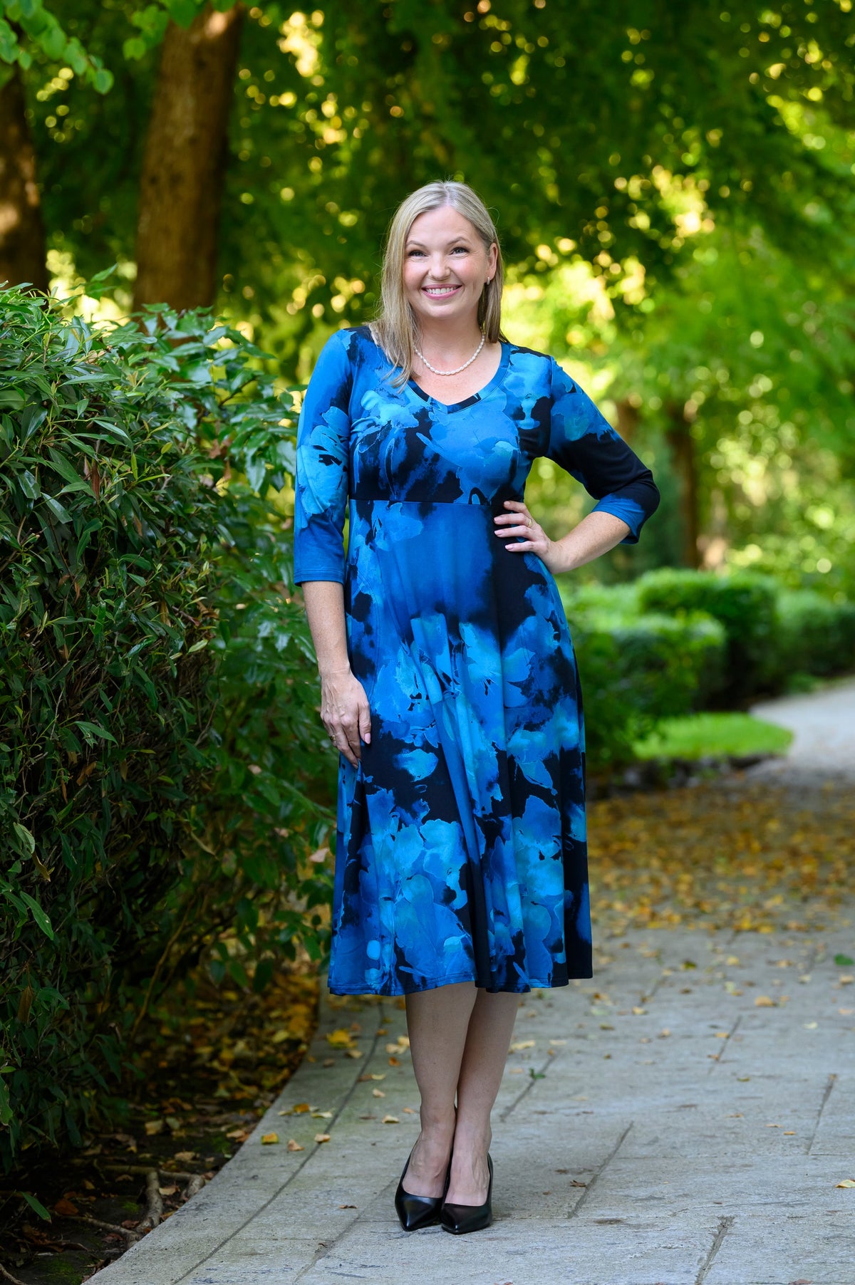 female model stands on a paved path in a park, hand on hip, wearing a blue and black floral midi dress with three-quarter sleeves and black heels.