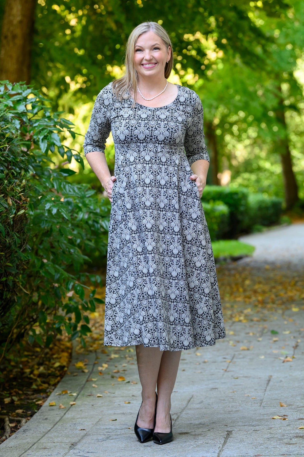 Front view of female model standing on a paved path in a wooded outdoor setting, wearing a knee-length gray midi dress with a black floral pattern, elbow-length sleeves, and black heels.