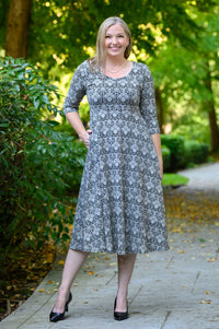 Front view of female model wearing a gray and white patterned midi dress with three-quarter sleeves, hand on hip, standing on a paved path in a park with lush green trees.