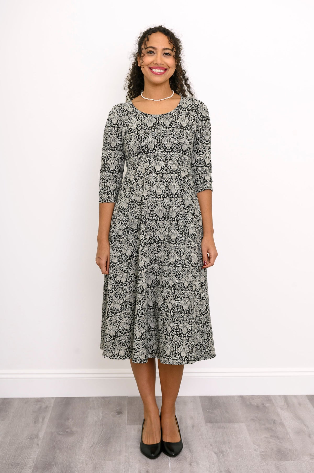 Front view of a female model wearing an everyday grey floral print dress with a scoop neck and three-quarter sleeves, with black heels, against white wall and gray floor