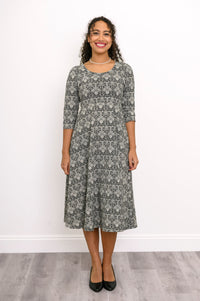 Front view of a female model wearing an everyday grey floral print dress with a scoop neck and three-quarter sleeves, with black heels, against white wall and gray floor
