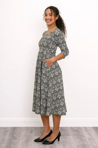 Side angled front view of a female model wearing a dark grey floral midi dress with black heels, against white wall and gray floor