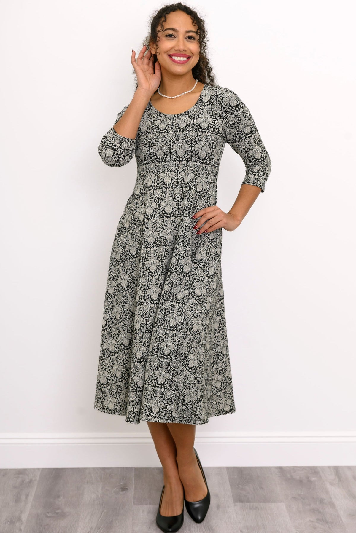 Front view of female model wearing a patterned dress with black heels against a white wall and gray floor
