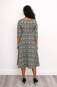 Back view of a female model wearing a grey patterned midi dress and black heels, against white wall and gray floor