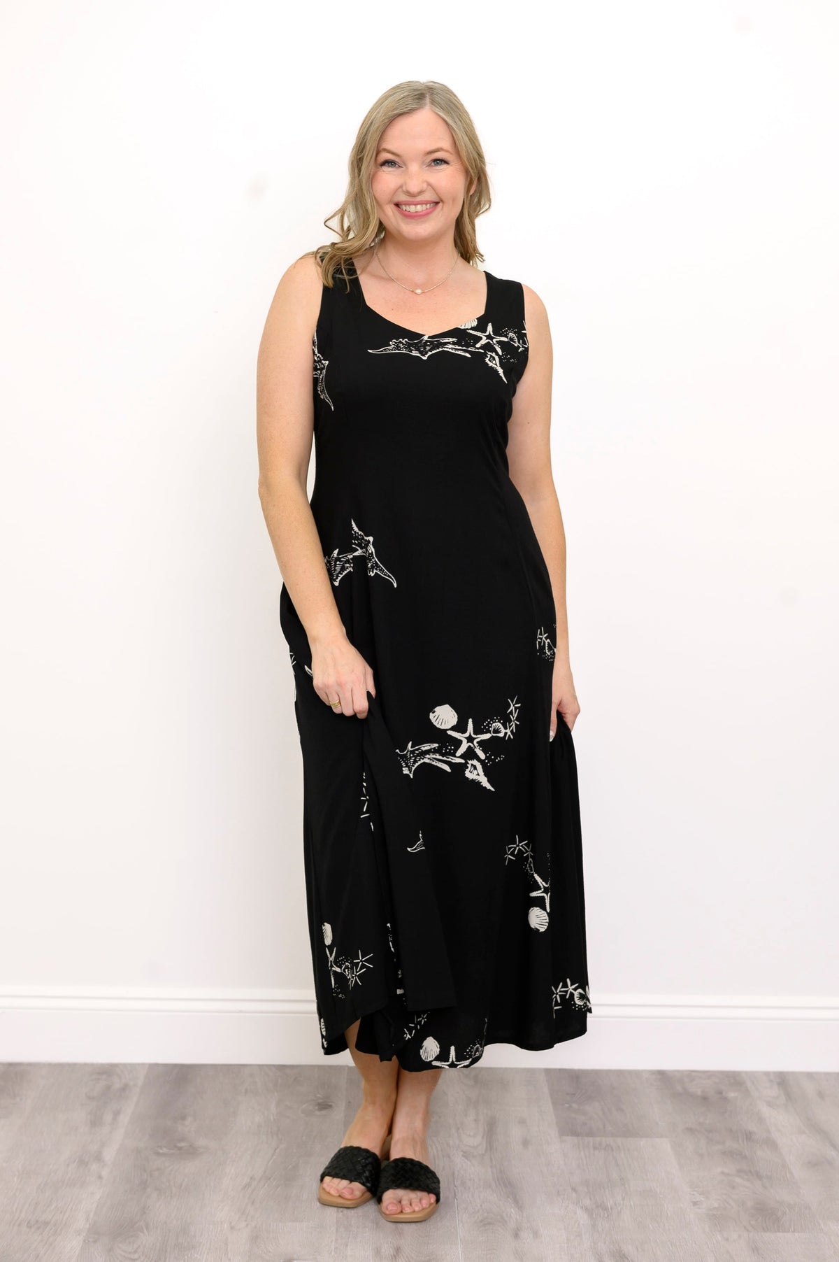 Woman wearing a long black sleeveless dress with white seashells and sandals against a white background, front view