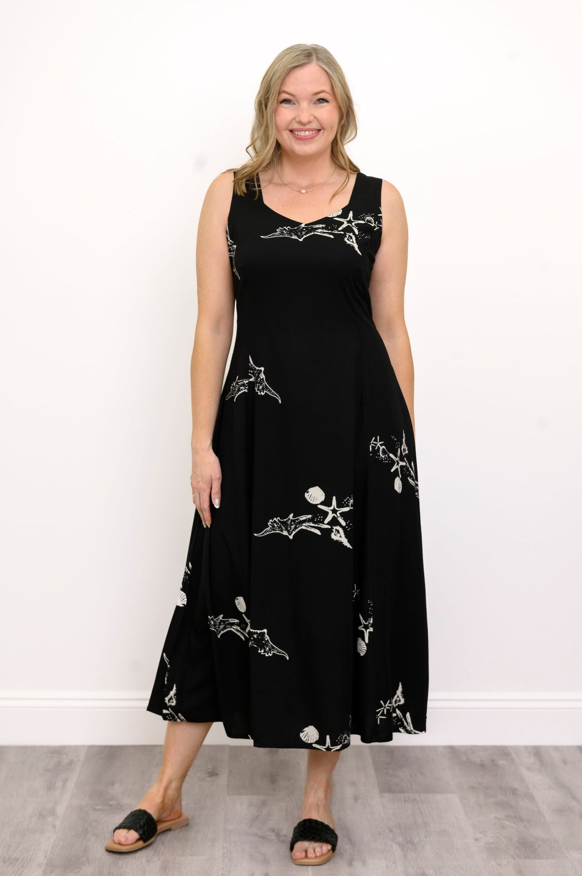 Woman wearing a long black sleeveless dress with white seashells and sandals against a white background, front view