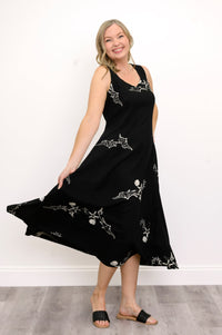 Woman wearing a long black sleeveless dress with white seashells and sandals against a white background, front view
