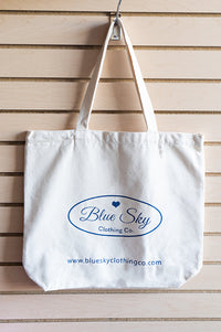 A white canvas tote bag with blue printed branding hanging from a metal hook on a light wood slatwall. The bag features the 'Blue Sky Clothing Co.