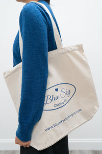 A person wearing a blue sweater carries a large off-white canvas tote bag with blue printed branding that reads Blue Sky Clothing Co.