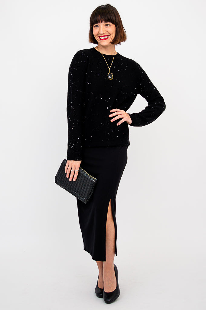 Full length front shot of a woman in a sparkly black knit sweater, black skirt, and leather handbag.