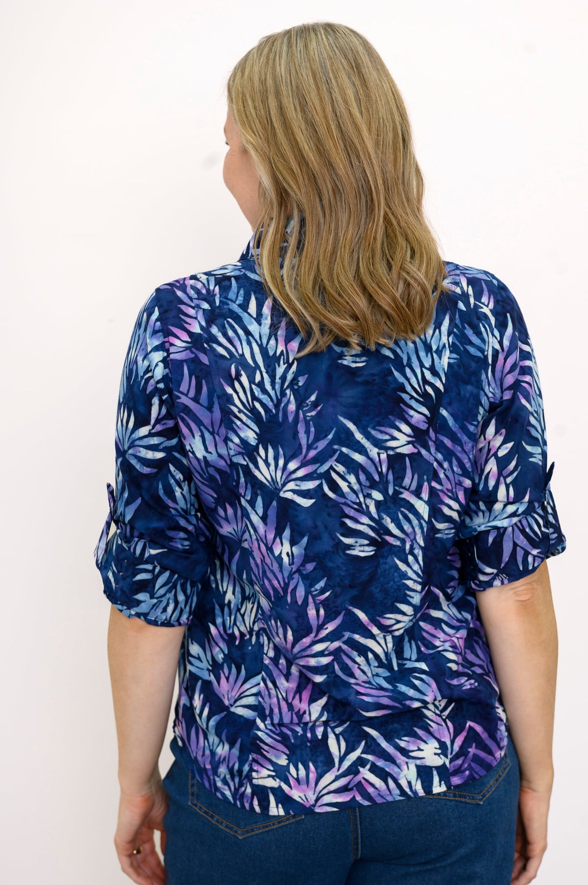 Back view of woman wearing a blue and purple patterned blouse and jeans against a white background