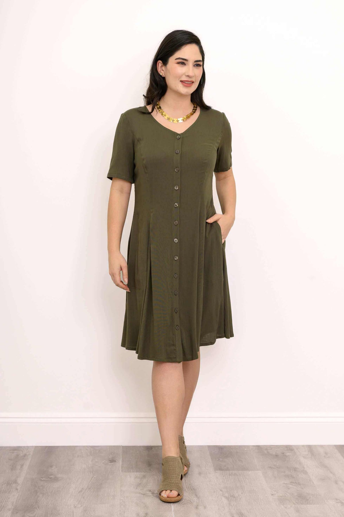 Woman with necklace wearing olive button-front dress, hands in pockets